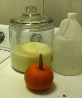 Image shows a large glass jar on a washing machine with homemade laundry detergent in it, next to a jug of vinegar and a small pumpkin.