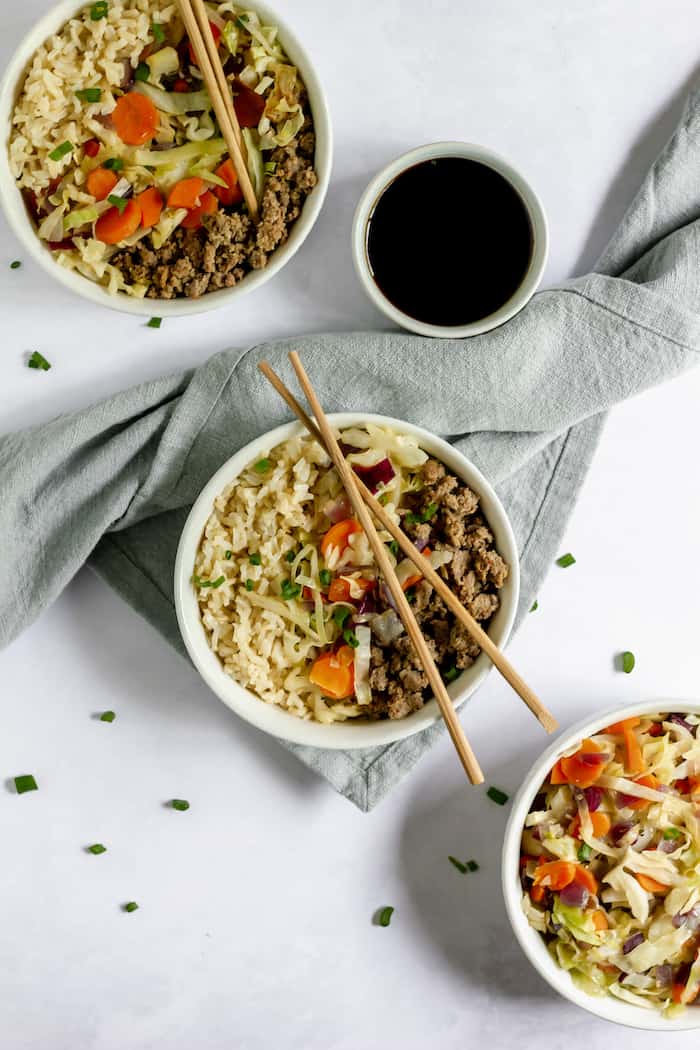 Image, taken from above, shows a couple of large bowls of egg roll in a bowl with veggies, ground meat and rice with chop sticks resting on the bowl. A smaller bowl of soy sauce sits nearby as well.