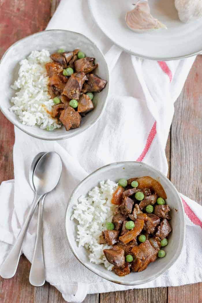 Image, taken from above, shows two bowls on a white towel on a table. The bowls are filled with white rice, as well as peas, onions, and beef heart. Two spoons sit next to the bowls.