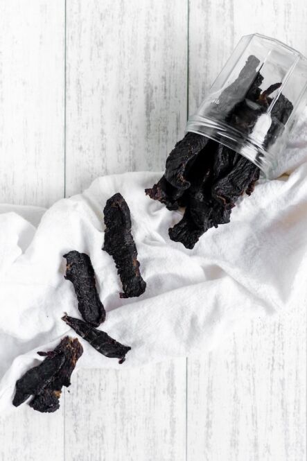Image, taken from above, shows a small jar of deer jerky on it's side, with several pieces of jerky spilled out onto a white towel.